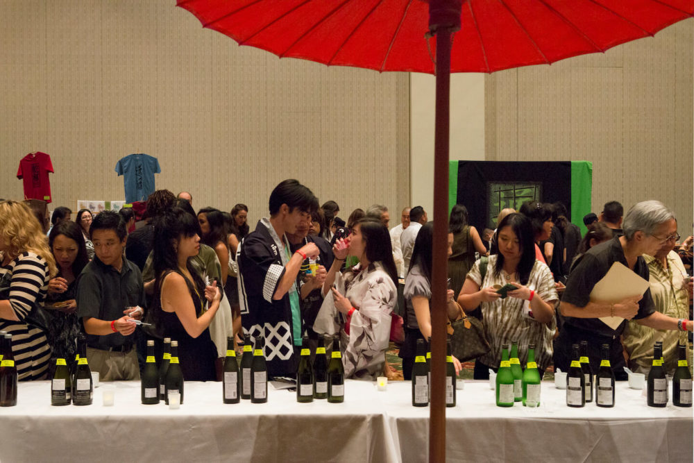 jos13hi024 A couple facing each other and sampling their sake in the crowd behind sake display table (2013 Honolulu)