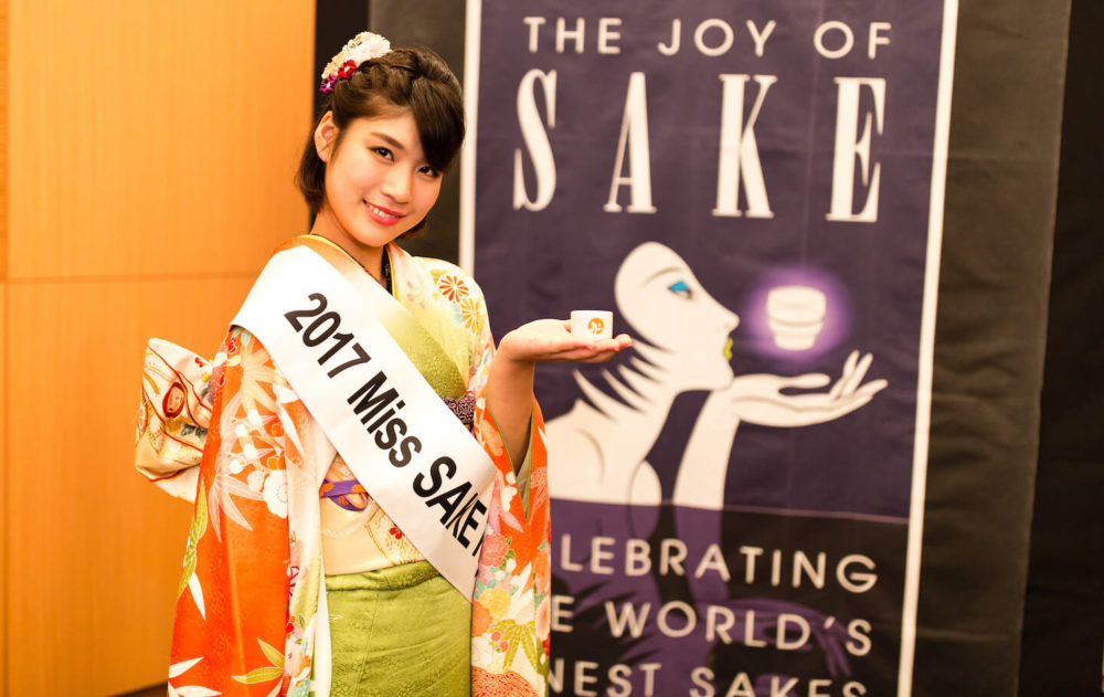jos16tk285 Miss Sake of 2017 holding a sake cup in front of the Joy of Sake banner (2016 Tokyo)